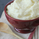 A red bowl filled with creamy mashed potatoes sits on a folded napkin, next to a wooden spoon on a dark surface.