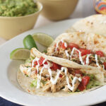 Two chicken tacos topped with diced tomatoes, lettuce, and a drizzle of white sauce are served on a white plate with lime wedges. Bowls of guacamole and salsa are blurred in the background.