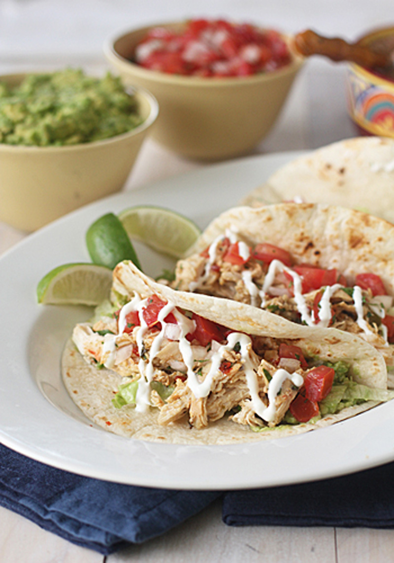 Two chicken tacos topped with diced tomatoes, lettuce, and a drizzle of white sauce are served on a white plate with lime wedges. Bowls of guacamole and salsa are blurred in the background.