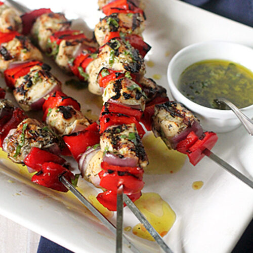 Grilled chicken and red bell pepper skewers on a white plate, served with a small bowl of green herb sauce, with a blue napkin underneath the plate.