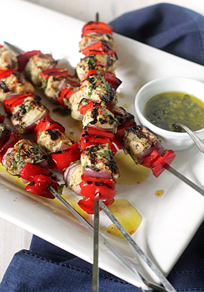 Grilled chicken and red bell pepper skewers on a white plate, served with a small bowl of green herb sauce, with a blue napkin underneath the plate.