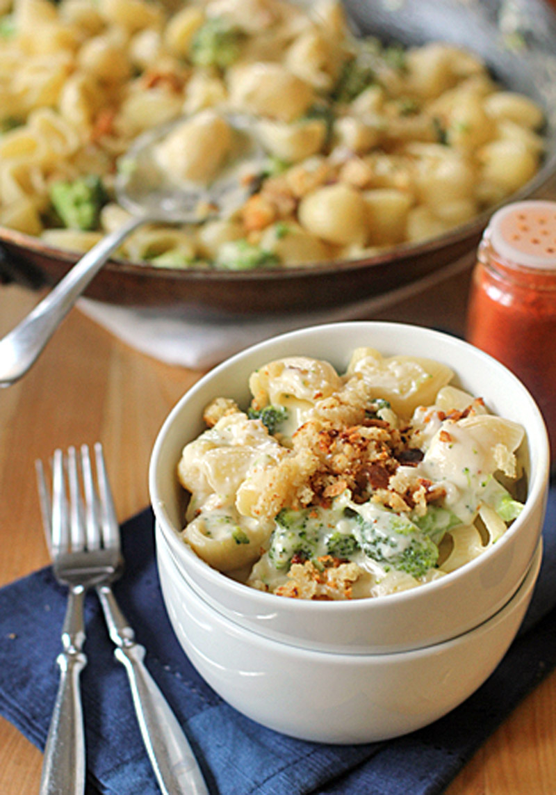 A bowl of pasta shells with broccoli in a creamy sauce, topped with crispy breadcrumbs, sits on a blue napkin with two forks. A skillet with more pasta and a shaker of red spice are in the background.