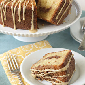 A slice of marbled bundt cake with glaze sits on a white plate with a fork, in front of the full cake on a stand. The scene includes a yellow patterned napkin and utensils on a light blue tablecloth.