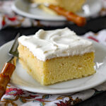 A slice of yellow cake with white frosting on a white plate, placed on a patterned napkin, with a fork beside it. Another plate with a similar slice is seen in the background.
