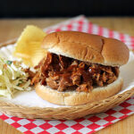 A pulled pork sandwich with barbecue sauce sits on a white plate, accompanied by coleslaw and a potato chip, on a wicker tray with a red and white checkered napkin underneath.