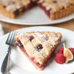 A slice of berry pie with a lattice crust is served on a white plate with a fork, garnished with fresh raspberries and a fig. The pie is dusted with powdered sugar and the rest of the pie is visible in the background.