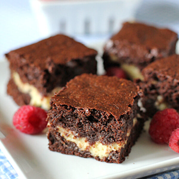 Four chocolate brownies with a creamy filling are arranged on a white plate, garnished with fresh raspberries. In the background, a basket of more raspberries is visible, all set atop a blue checkered cloth.