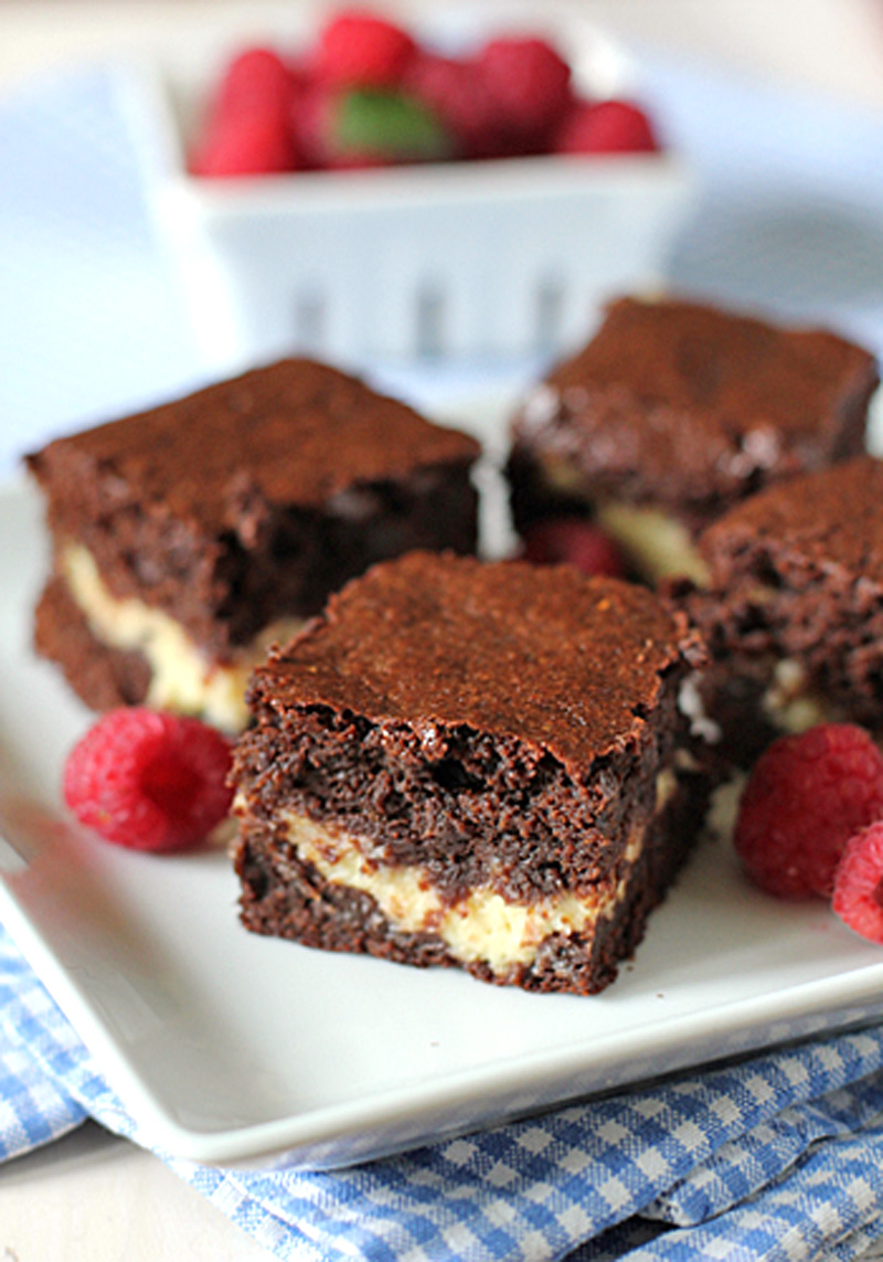 Four chocolate brownies with a creamy filling are arranged on a white plate, garnished with fresh raspberries. In the background, a basket of more raspberries is visible, all set atop a blue checkered cloth.