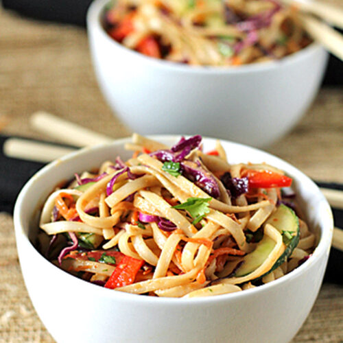 Two white bowls filled with colorful noodle salad, including vegetables like red bell pepper, purple cabbage, and zucchini, are placed on a woven surface with chopsticks resting nearby.