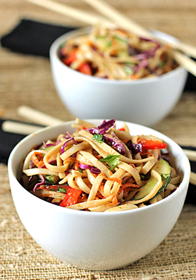Two white bowls filled with colorful noodle salad, including vegetables like red bell pepper, purple cabbage, and zucchini, are placed on a woven surface with chopsticks resting nearby.