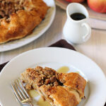 A slice of apple crumble pie on a white plate with a fork, drizzled with syrup. In the background, a whole pie, a small pitcher of syrup, and a bowl of apples are visible.
