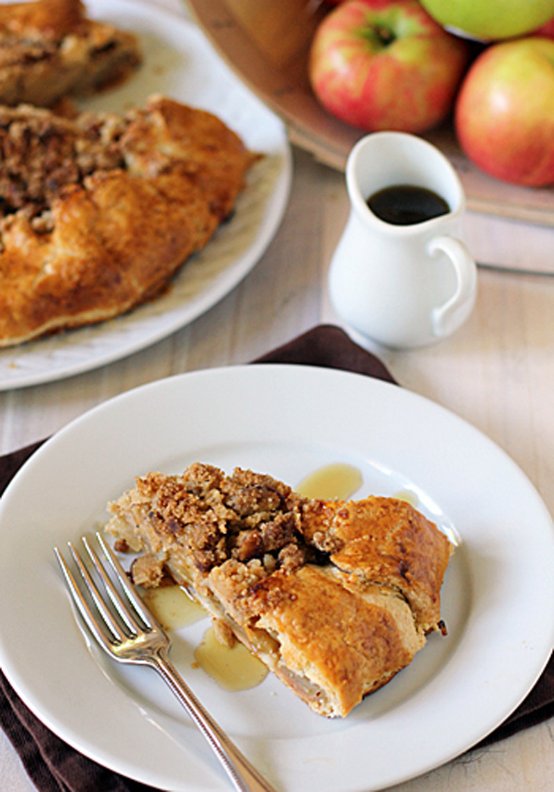 A slice of apple crumble pie on a white plate with a fork, drizzled with syrup. In the background, a whole pie, a small pitcher of syrup, and a bowl of apples are visible.