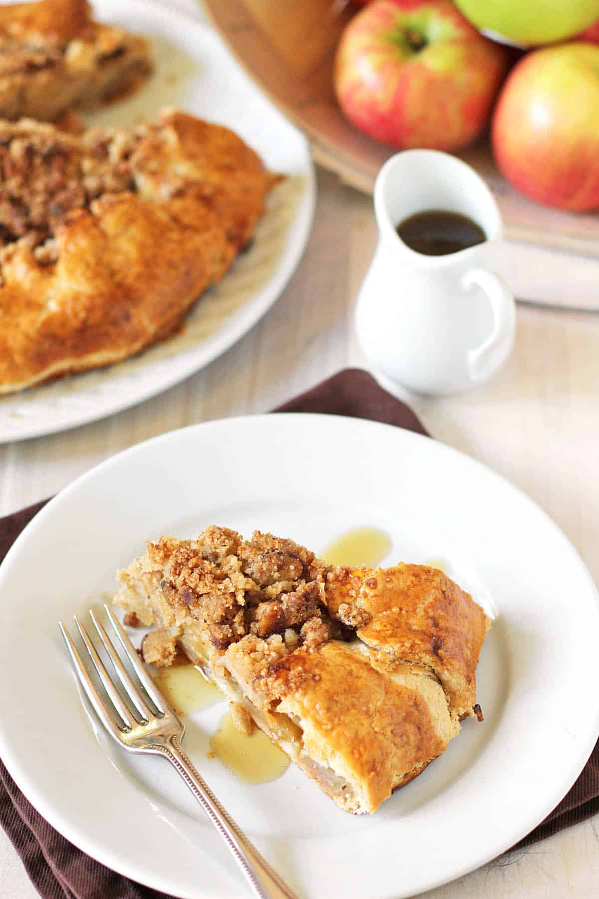 A slice of apple pie with crumb topping sits on a white plate beside a fork, with syrup drizzled on top. A whole pie, a jug of syrup, and fresh apples are in the background.