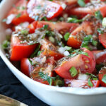 A white bowl filled with a fresh tomato salad, featuring chopped tomatoes, diced onions, green herbs, and a vinaigrette dressing, placed on a dark cloth next to a silver serving spoon.
