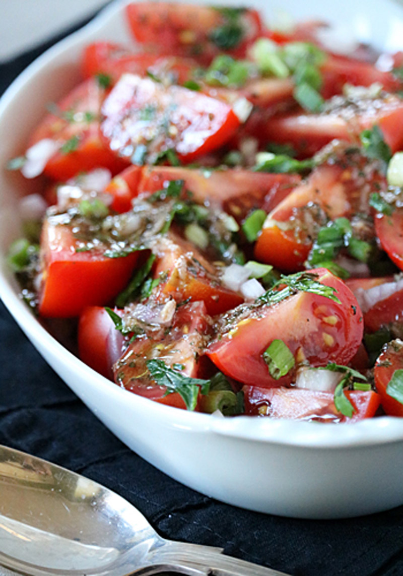 A white bowl filled with a fresh tomato salad, featuring chopped tomatoes, diced onions, green herbs, and a vinaigrette dressing, placed on a dark cloth next to a silver serving spoon.