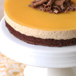 A layered mousse cake with a glossy caramel top, chocolate mousse middle, and brownie base, garnished with chocolate shavings, displayed on a white cake stand.