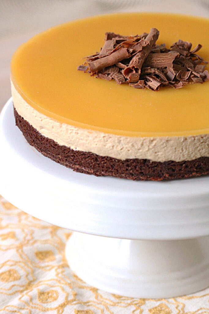 A layered mousse cake with a glossy caramel top, chocolate mousse middle, and brownie base, garnished with chocolate shavings, displayed on a white cake stand.