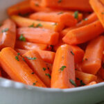 A white dish filled with cooked carrot sticks garnished with chopped herbs, placed on a green textured surface.