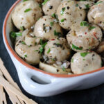 A white dish filled with marinated mushrooms garnished with chopped herbs and onions, placed on a dark surface with a few wooden toothpicks beside the dish.