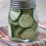 A glass mason jar filled with sliced cucumbers and herbs in liquid, sitting on a striped cloth on a light wooden surface.