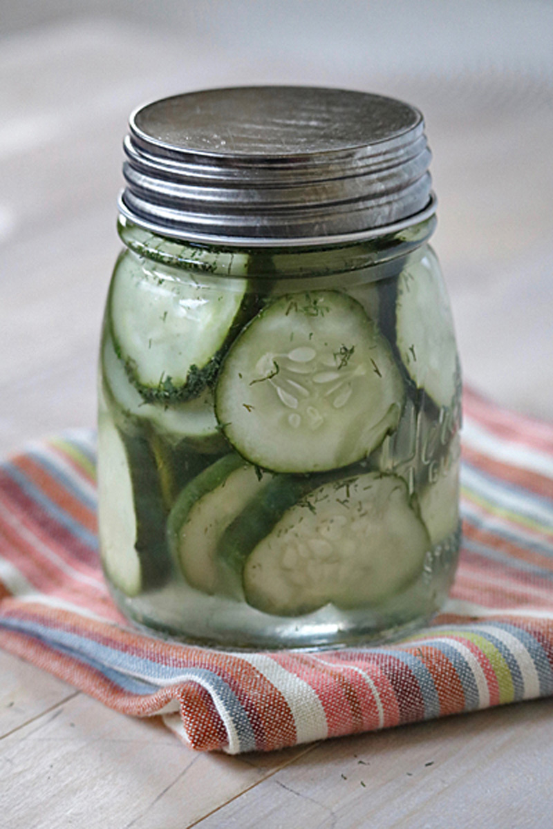 A glass mason jar filled with sliced cucumbers and herbs in liquid, sitting on a striped cloth on a light wooden surface.