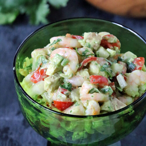A green glass dish filled with shrimp ceviche, containing shrimp, avocado, tomato, cucumber, and cilantro, sits on a dark surface with a lime and tortilla chips in the background.