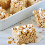 A peanut butter rice krispie treat topped with mini marshmallows and a drizzle of peanut butter sits on parchment paper, with more treats visible in the background in a white dish.