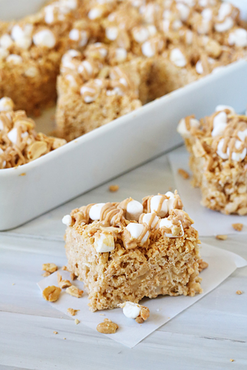 A peanut butter rice krispie treat topped with mini marshmallows and a drizzle of peanut butter sits on parchment paper, with more treats visible in the background in a white dish.
