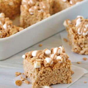 A close-up of Fluffer-nutter Treats—peanut butter Rice Krispies treats topped with mini marshmallows and a drizzle of peanut butter, with more treats in a white dish behind.