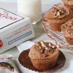 A chocolate muffin with almond pieces sits on an unwrapped liner next to a packaged Goo Goo Cluster candy, a glass of milk with a red-striped straw, a box labeled Made in Nashville, and more muffins on a wire rack.