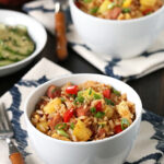 Two white bowls filled with colorful fried rice, featuring pieces of pineapple, diced red bell pepper, green onions, and bits of meat, are set on patterned napkins next to a fork and a side dish.