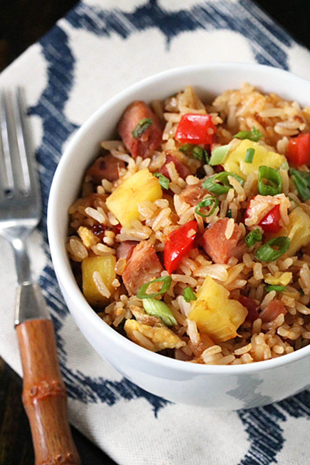 A white bowl filled with fried rice, diced ham, pineapple chunks, red bell peppers, and green onions, placed on a patterned cloth napkin next to a fork with a wooden handle.