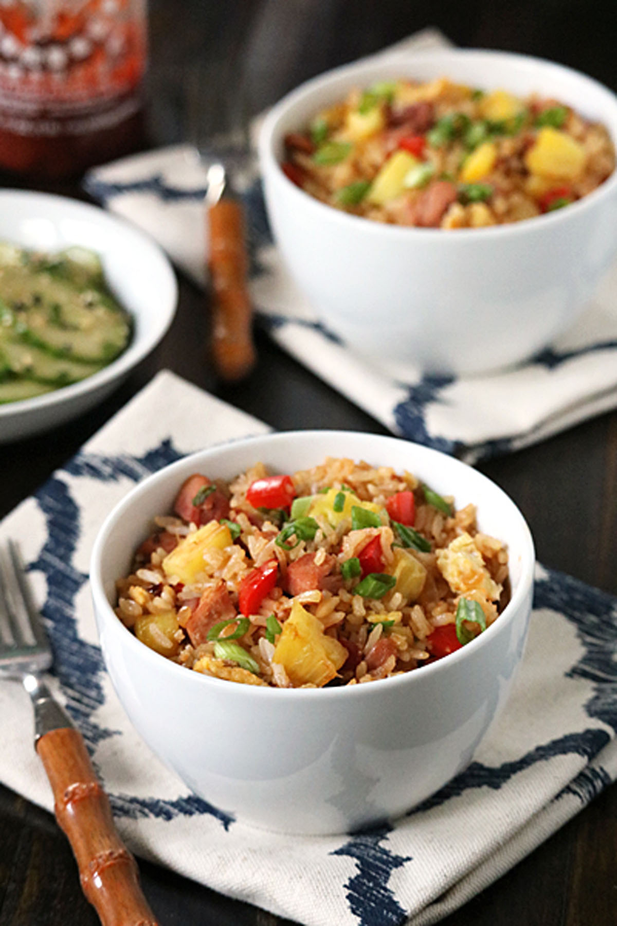Two white bowls filled with fried rice featuring pineapple chunks, ham, red bell pepper, and green onions, set on patterned napkins beside forks, with a small side dish and a bottle in the background.