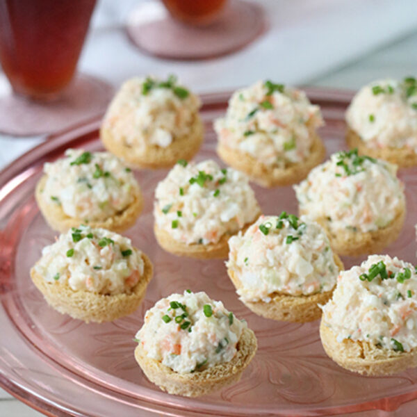 Round crackers topped with a creamy salad mixture, garnished with chopped chives, arranged on a pink glass platter. In the background, two glasses of a dark beverage sit on a white napkin.