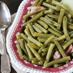 A vintage red patterned plate filled with cooked green beans and pieces of ham, placed on a white plate with a beige napkin and a silver spoon beside it.
