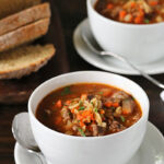Two white bowls of hearty vegetable and beef soup sit on saucers, garnished with herbs. In the background, slices of rustic bread are arranged on a wooden platter. The setting is a dark wooden table.