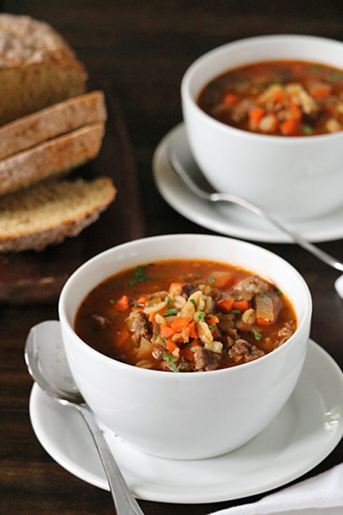 Two white bowls of hearty vegetable and beef soup sit on saucers, garnished with herbs. In the background, slices of rustic bread are arranged on a wooden platter. The setting is a dark wooden table.