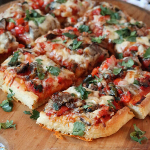 A rectangular pizza topped with melted cheese, vegetables, and fresh cilantro, sliced into squares and served on a wooden board. A colorful napkin is placed nearby.