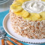 A round cake covered with toasted coconut flakes and decorated with a ring of pineapple slices and white frosting, placed on a white plate with wooden-handled forks beside it.