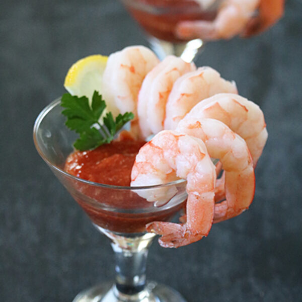Two cocktail glasses filled with shrimp cocktail, featuring large shrimp hooked on the rim, cocktail sauce inside, and garnished with parsley and a lemon wedge. The background is a dark, blurred surface.