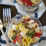 A glass filled with rotini pasta salad, featuring sliced peppers, mozzarella balls, salami, artichokes, and herbs. Another glass with the same salad and two forks are visible in the background on a patterned napkin.