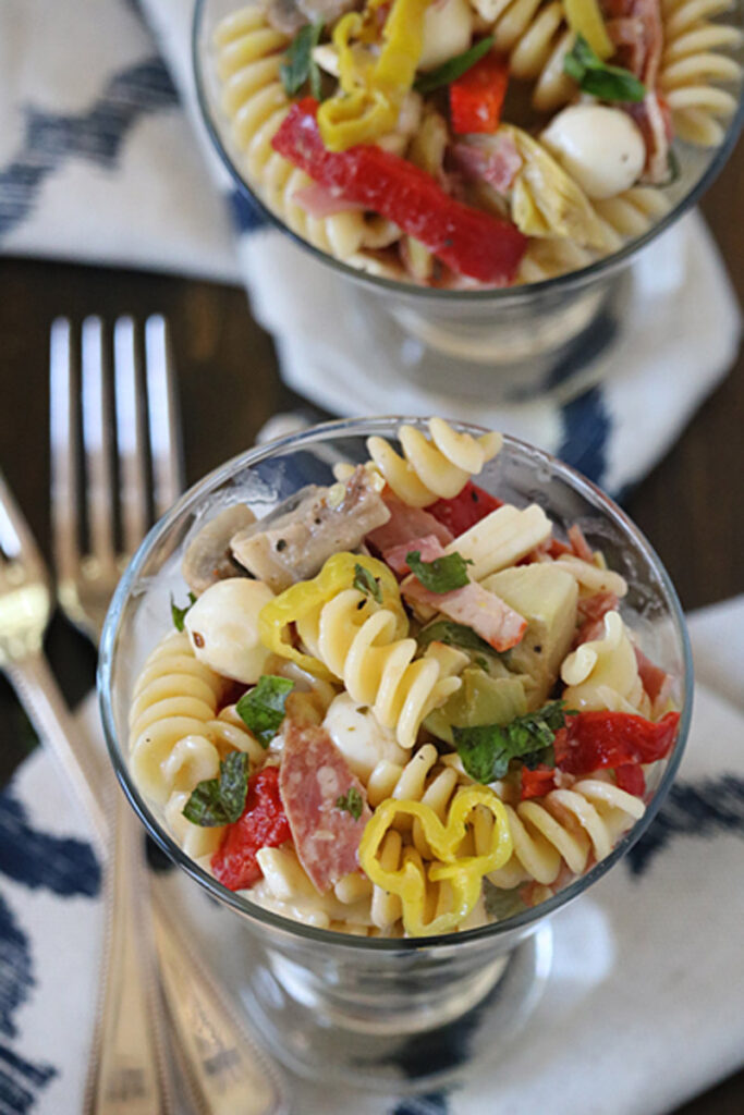 A glass filled with rotini pasta salad, featuring sliced peppers, mozzarella balls, salami, artichokes, and herbs. Another glass with the same salad and two forks are visible in the background on a patterned napkin.