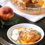 A bowl of peach cobbler topped with a scoop of vanilla ice cream is in the foreground, with a fork beside it. In the background, a peach and a glass baking dish of cobbler are on a dark wooden table.