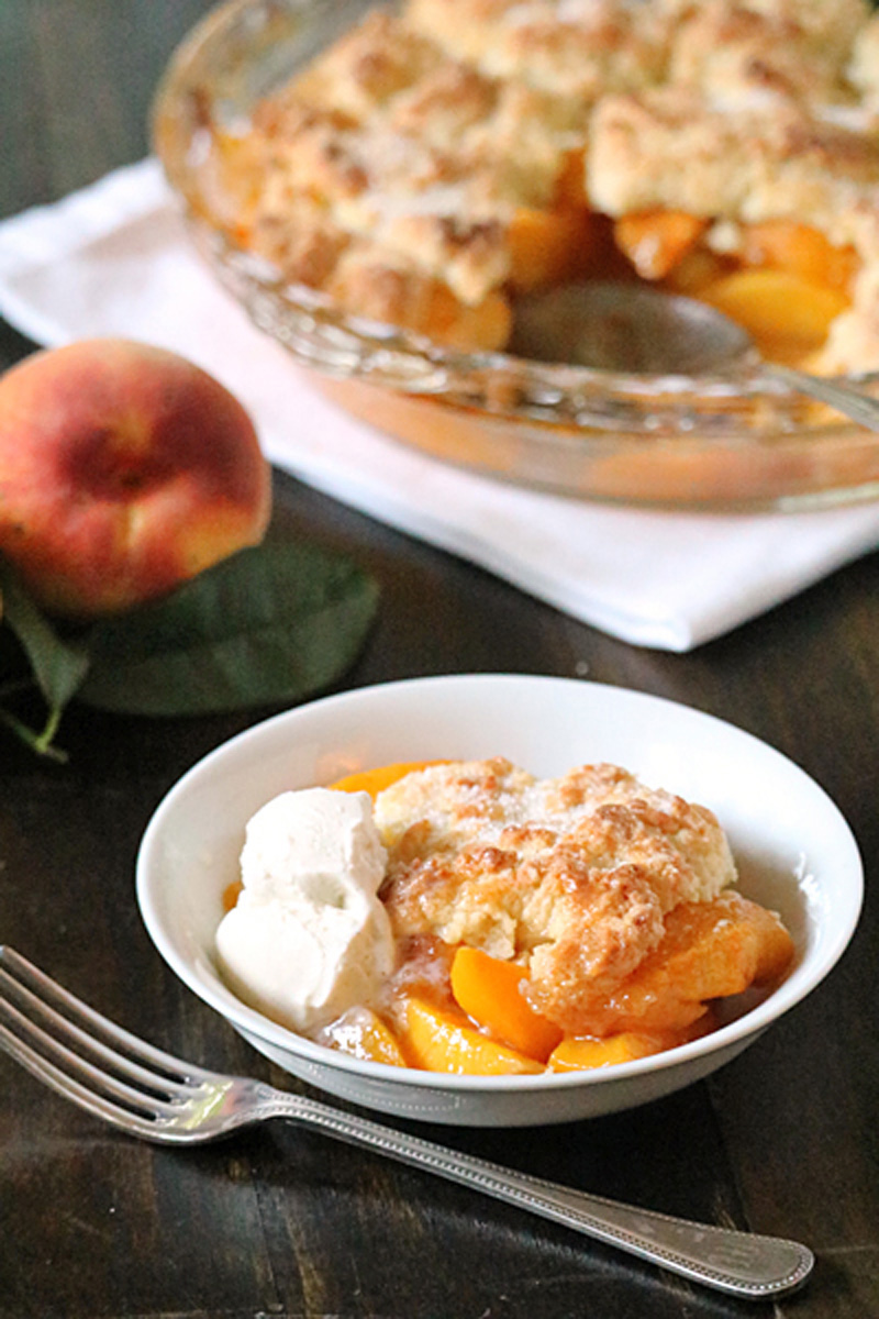 A bowl of peach cobbler topped with a scoop of vanilla ice cream is in the foreground, with a fork beside it. In the background, a peach and a glass baking dish of cobbler are on a dark wooden table.
