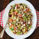 A white oval plate with chopped salad including pasta, lettuce, diced chicken, tomatoes, and vegetables, sits on a red and white checkered napkin with a serving spoon on a wooden table.