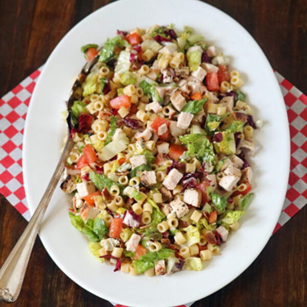 A white oval plate with chopped salad including pasta, lettuce, diced chicken, tomatoes, and vegetables, sits on a red and white checkered napkin with a serving spoon on a wooden table.