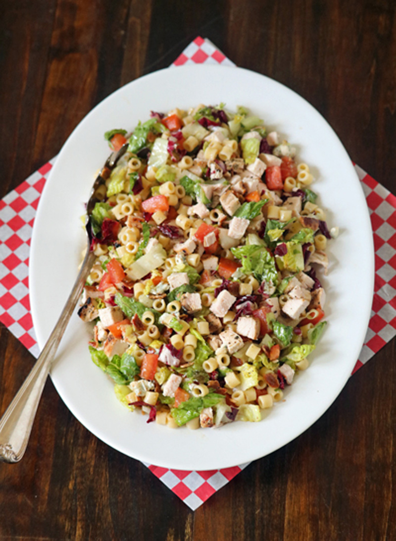 A white oval plate with chopped salad including pasta, lettuce, diced chicken, tomatoes, and vegetables, sits on a red and white checkered napkin with a serving spoon on a wooden table.