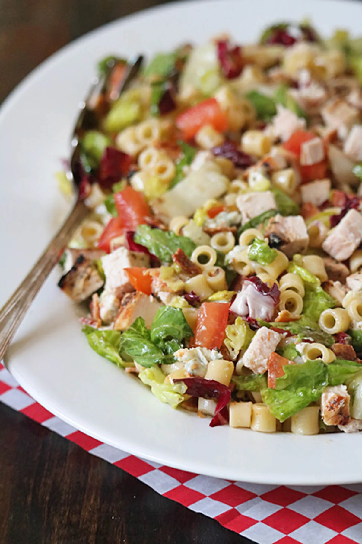 A white plate with Portillo's Chopped Salad featuring lettuce, diced tomatoes, grilled chicken, cheese cubes, pasta, and purple cabbage. A fork rests on the plate atop a red and white checkered cloth.