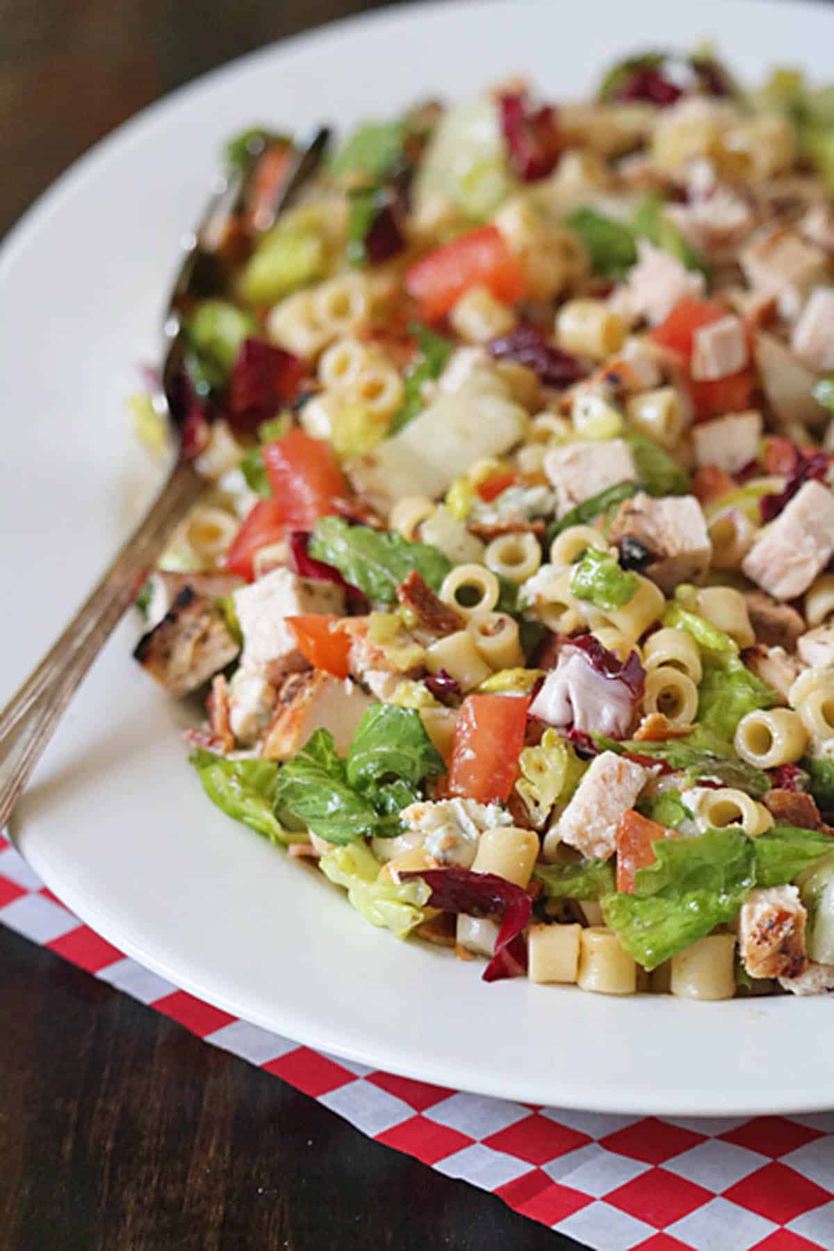 A white plate with Portillo's Chopped Salad featuring lettuce, diced tomatoes, grilled chicken, cheese cubes, pasta, and purple cabbage. A fork rests on the plate atop a red and white checkered cloth.