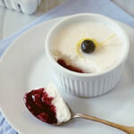 A ramekin of creamy dessert topped with a blueberry and lemon twist sits on a white plate. A spoonful of the dessert, showing a layer of berry filling, rests beside it. Fresh blueberries are in the background.
